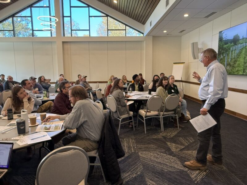 Presenter standing up in front of a high-ceilings meeting room with round tables filled with attendees sitting around them.