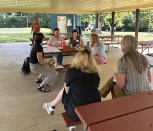 Group sitting around picnic tables under a shelter outside looking conversating, and looking at a large post it